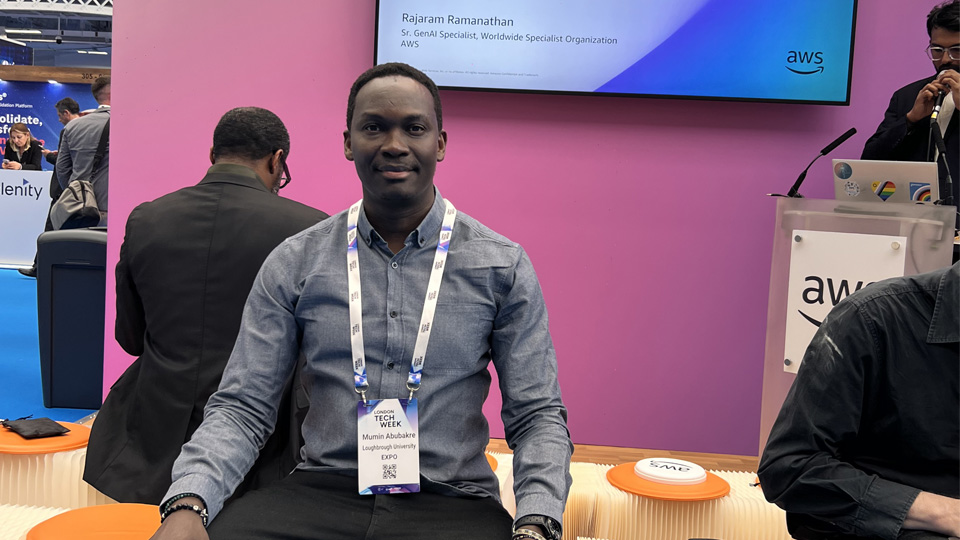 A man sits on an orange stool in front of a pink wall with a screen.